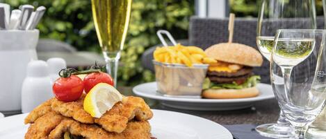 Una mesa de comedor con un plato de pescado rebozado, una hamburguesa, papas fritas y una copa de vino blanco.