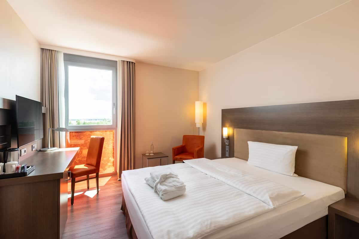 A hotel room with a bed, a desk with a TV, a chair, a lamp, and a view of the outdoors.