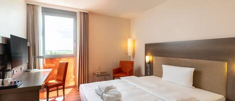 Une chambre d’hôtel avec un lit, un bureau avec une télévision, une chaise, une petite table et une fenêtre avec des rideaux.