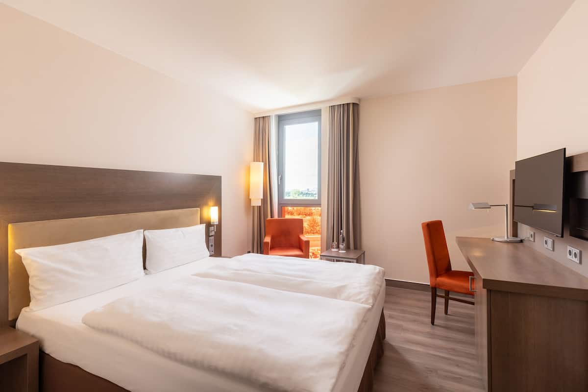 A hotel room with a large bed, a desk with a flat-screen TV, a chair, and a window with curtains.