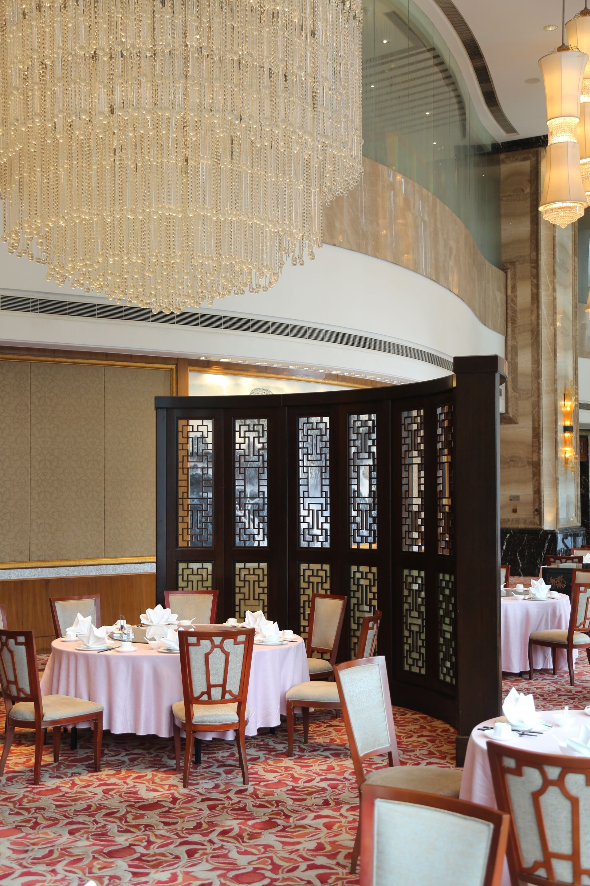 Un comedor con mesas redondas dispuestas para cuatro, cada una con manteles blancos y cubertería. Un gran candelabro ornamentado cuelga del techo y, al fondo, hay un biombo decorativo con diseños geométricos.