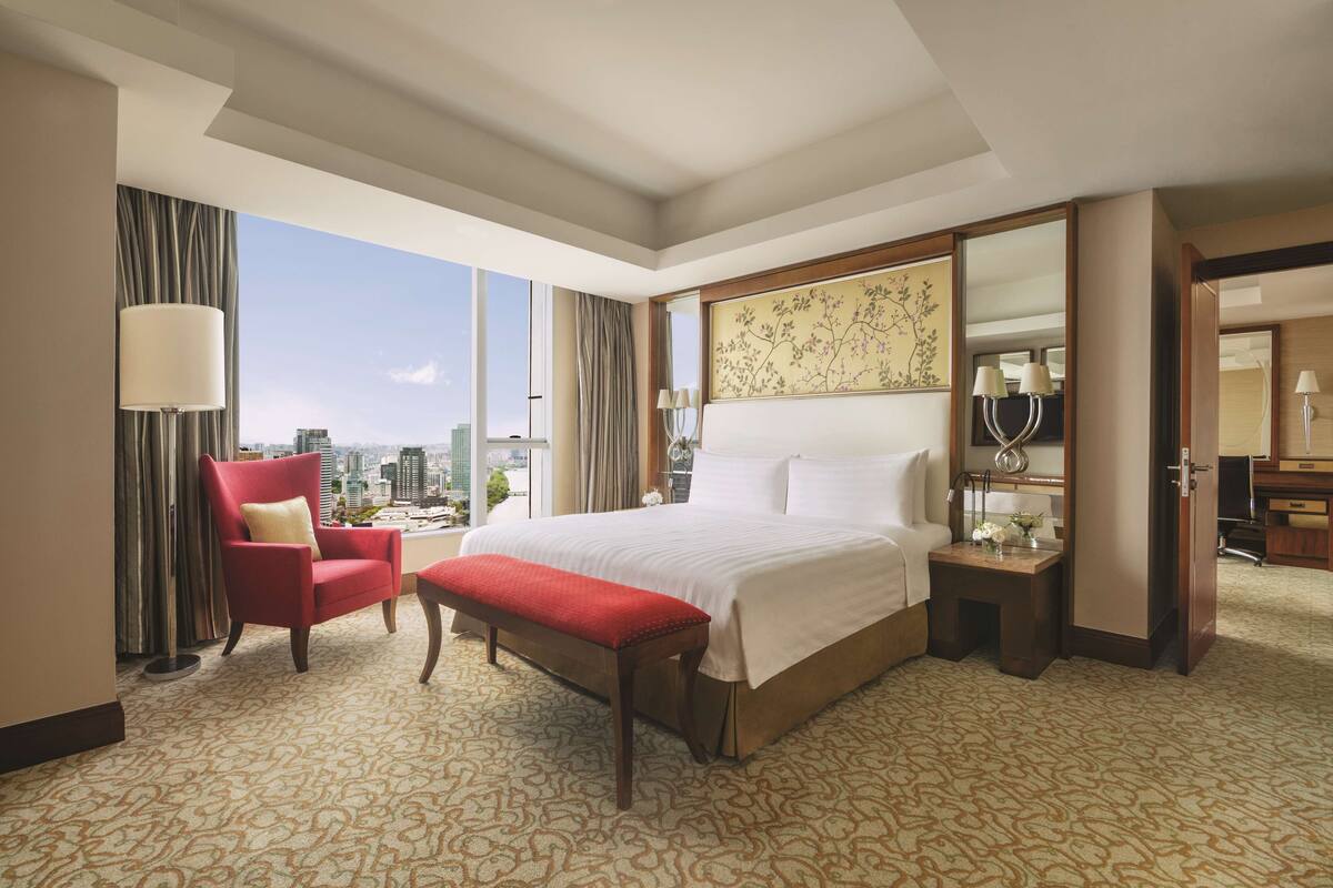 Habitación de hotel con una cama grande, un sillón rojo, un banco, una mesita de noche con una lámpara y vistas a la ciudad.