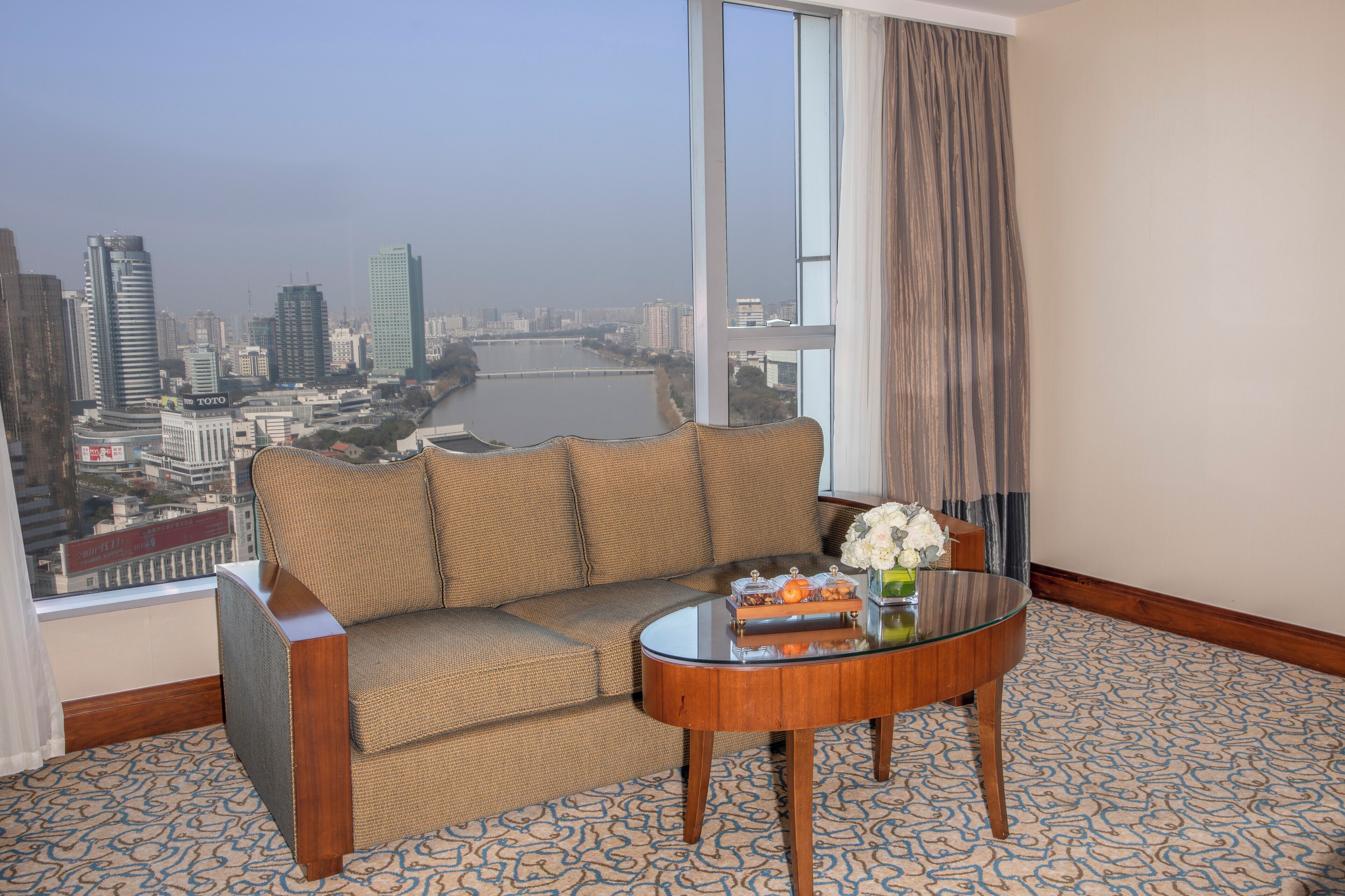 A hotel room with a sofa, a glass coffee table, and a view of a cityscape and river.