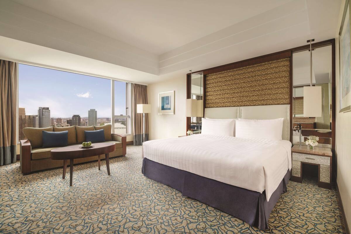 Habitación de hotel con una cama grande, un sofá, una mesita y vistas a la ciudad.