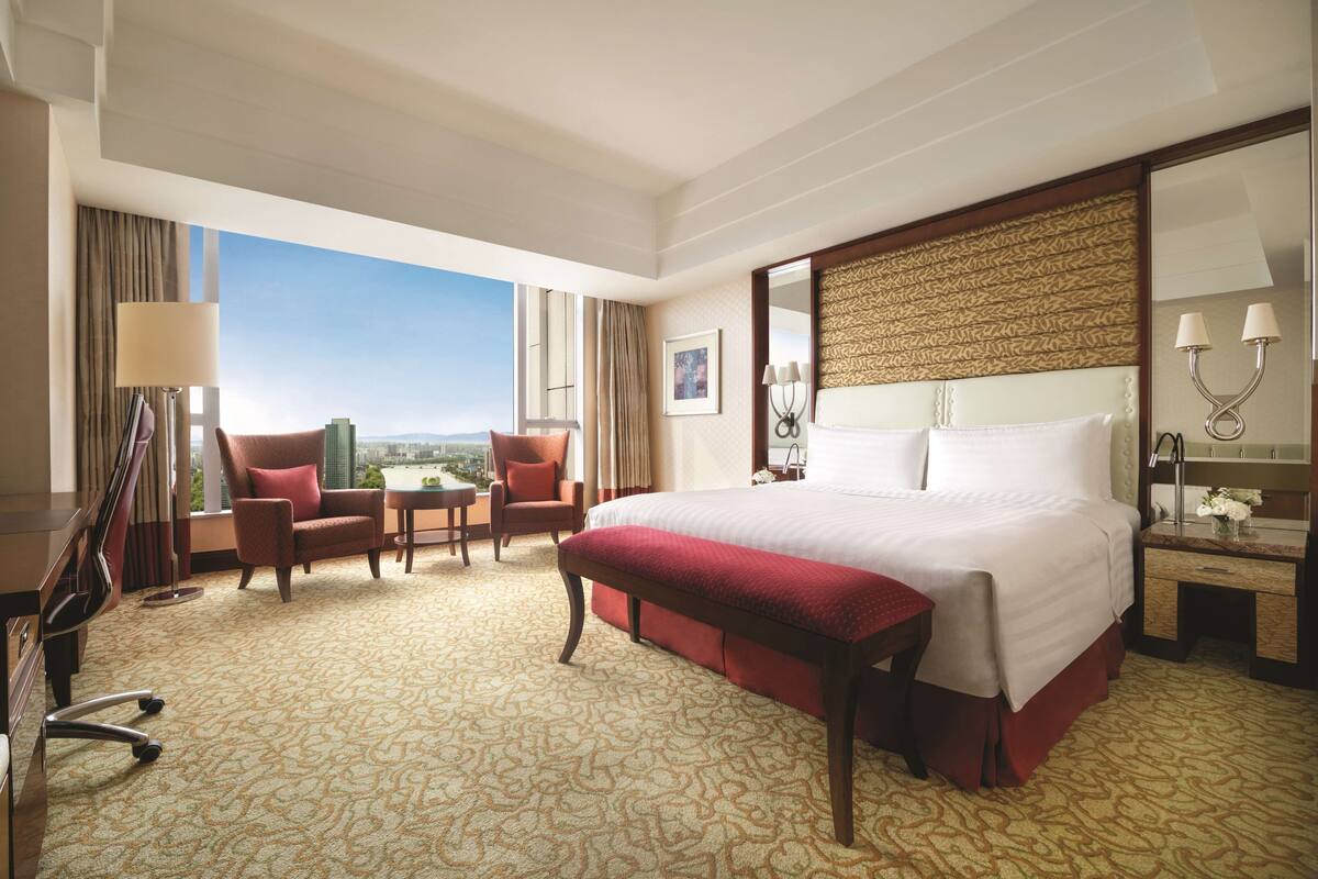 Habitación de hotel con una cama grande, dos sillones, un escritorio y vistas a la ciudad.