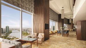 Un amplio vestíbulo de hotel con grandes ventanales que ofrecen vista a la ciudad, un área de estar con sofás y sillones, y un comedor con mesas y sillas.