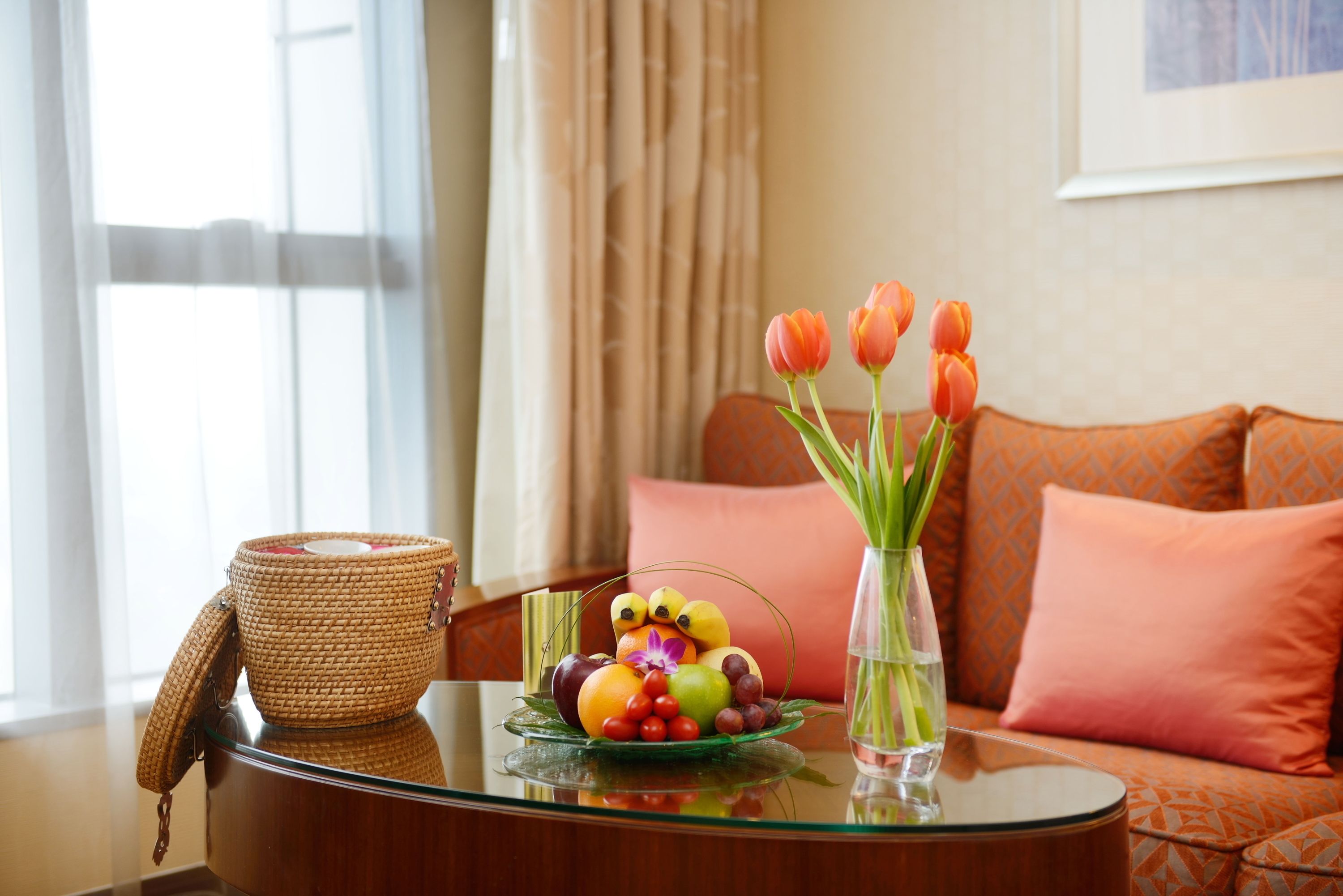 A glass table with a woven basket, a fruit bowl, and a vase with tulips.