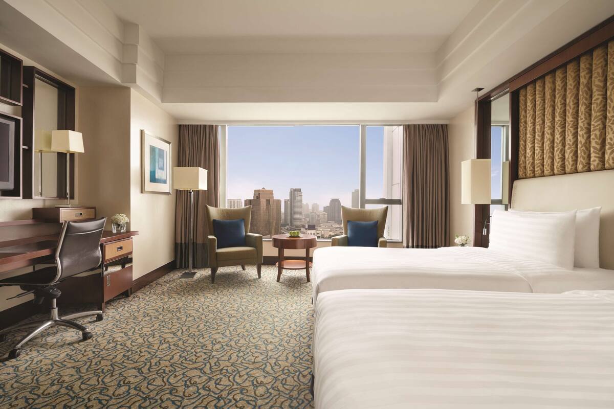 Habitación de hotel con una cama grande, un escritorio con una silla, una mesita con dos sillas y vistas a la silueta urbana a través de amplias ventanas.