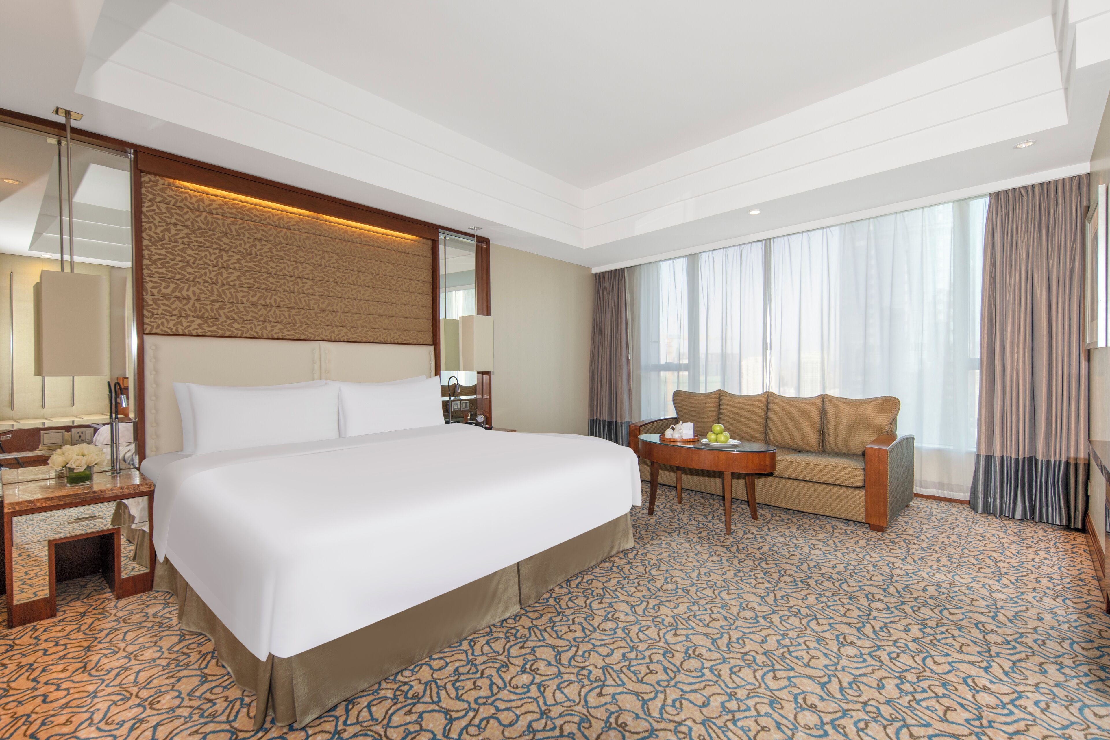 A hotel room with a large bed, a sofa, a small table with fruit, and a mirror.