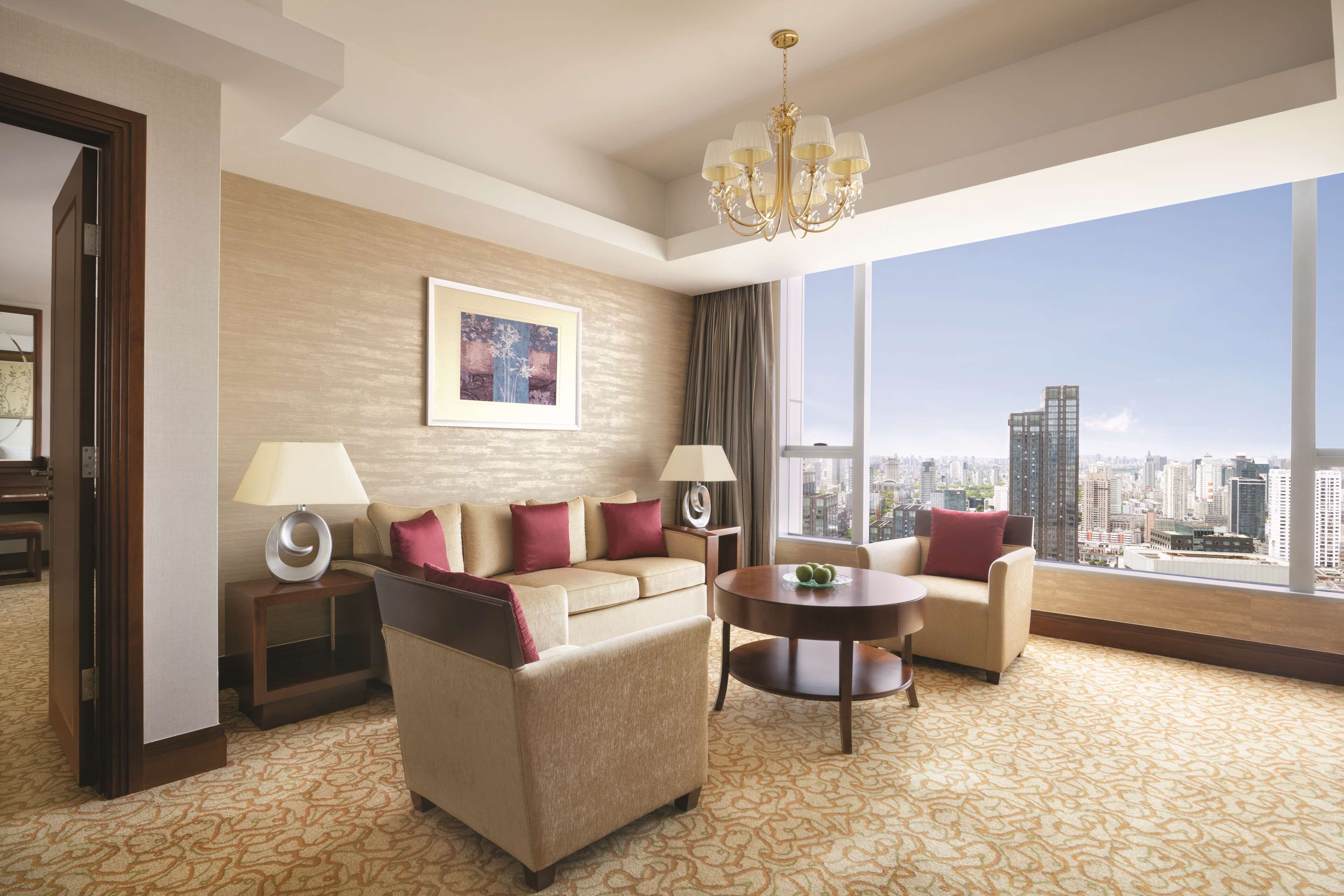 A hotel room with a sofa, armchairs, a coffee table, and a large window offering a city view.