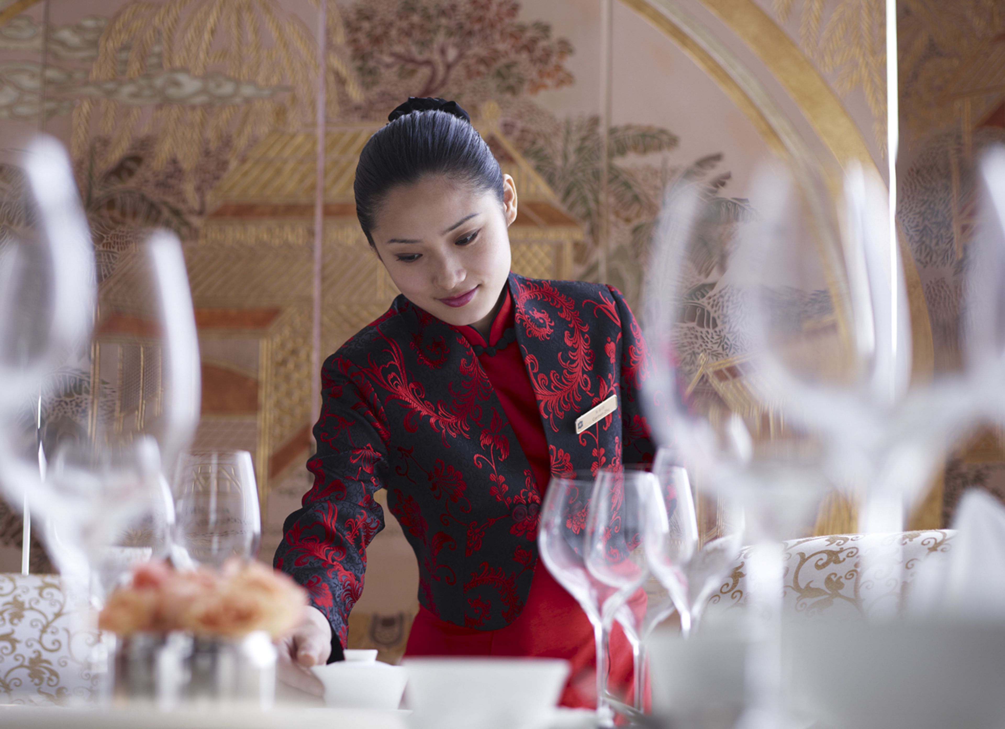 A person in a red and black uniform serving food at a table with a white tablecloth and gold patterns.