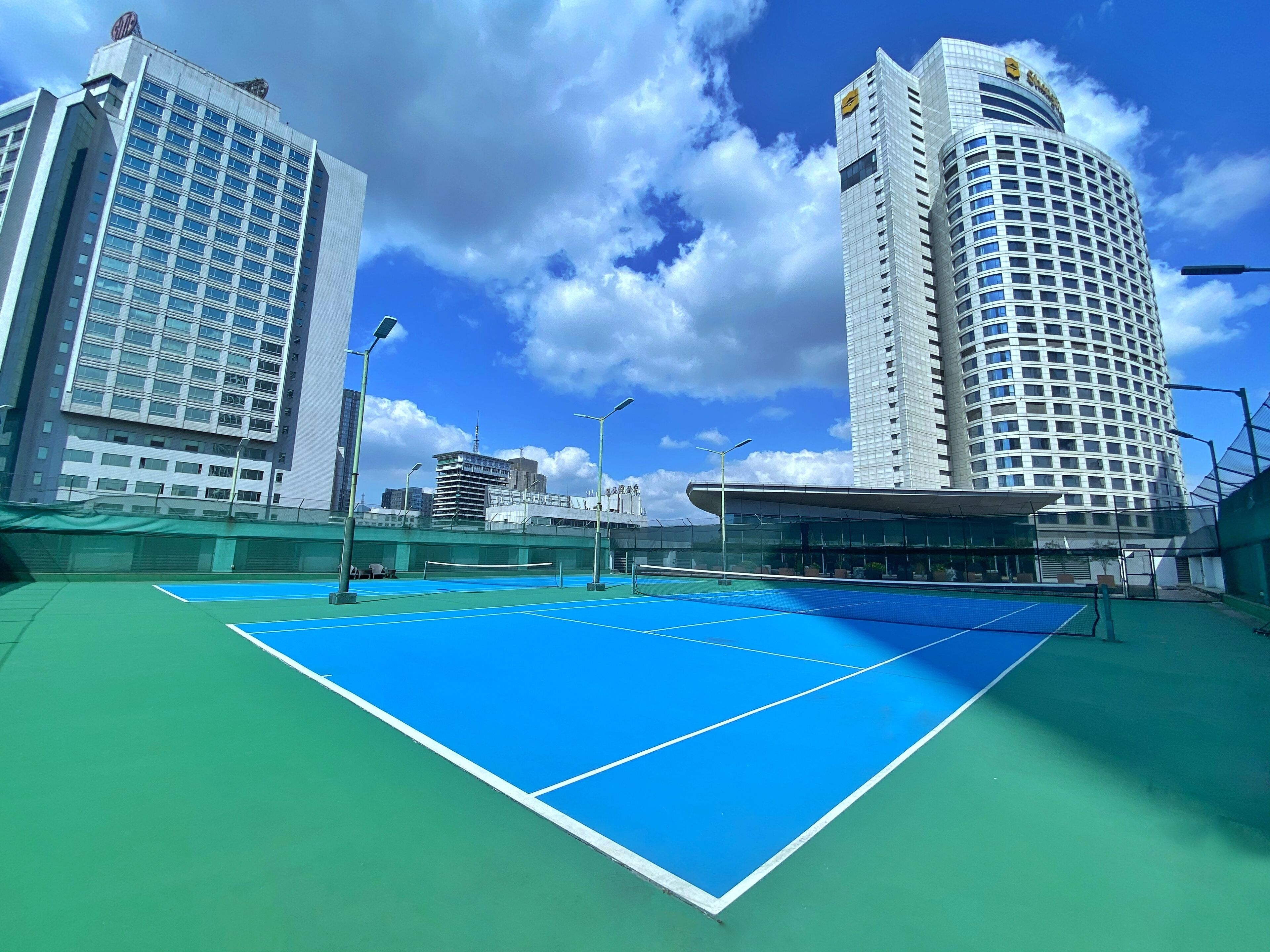 A tennis court with a blue surface, surrounded by modern buildings with glass facades and a clear sky.