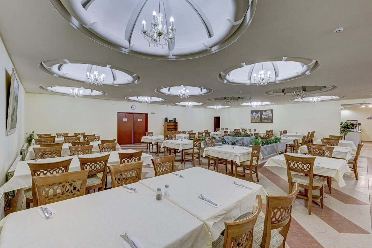 A large dining hall with multiple tables and chairs, a chandelier, and a clock on the wall.