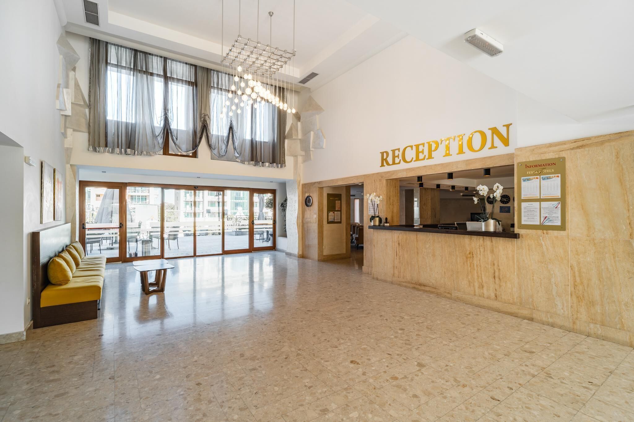 A hotel reception area with a reception desk, a seating area with a sofa and a table, and large windows allowing natural light.