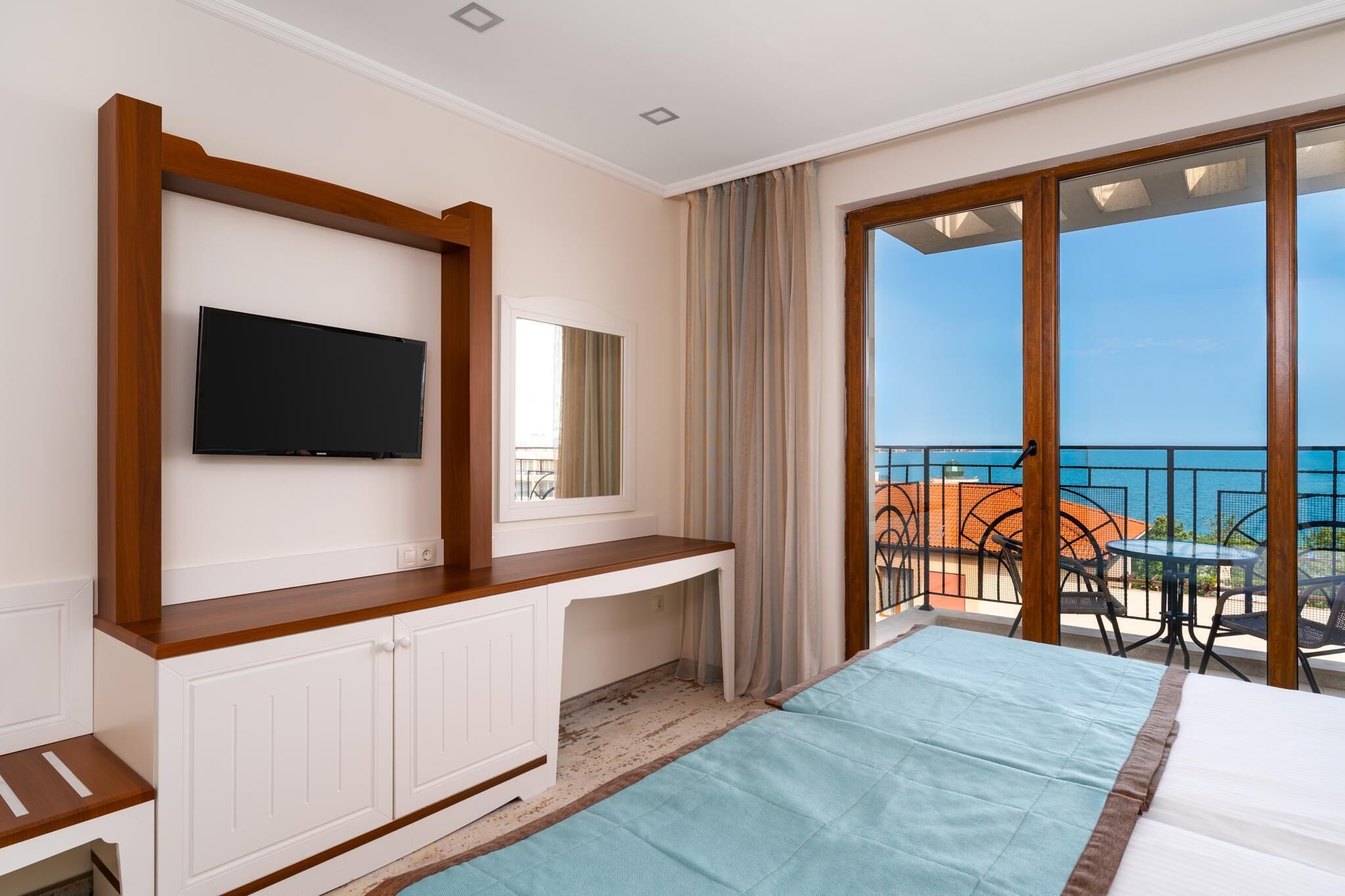 A hotel room with a bed, a TV mounted on a wooden cabinet, a balcony with outdoor seating, and a view of the sea.