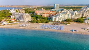 An aerial view of a coastal resort with multiple hotel buildings, a beach with sun loungers, and a clear view of the sea. - Festa Via Pontica (Pomorie)