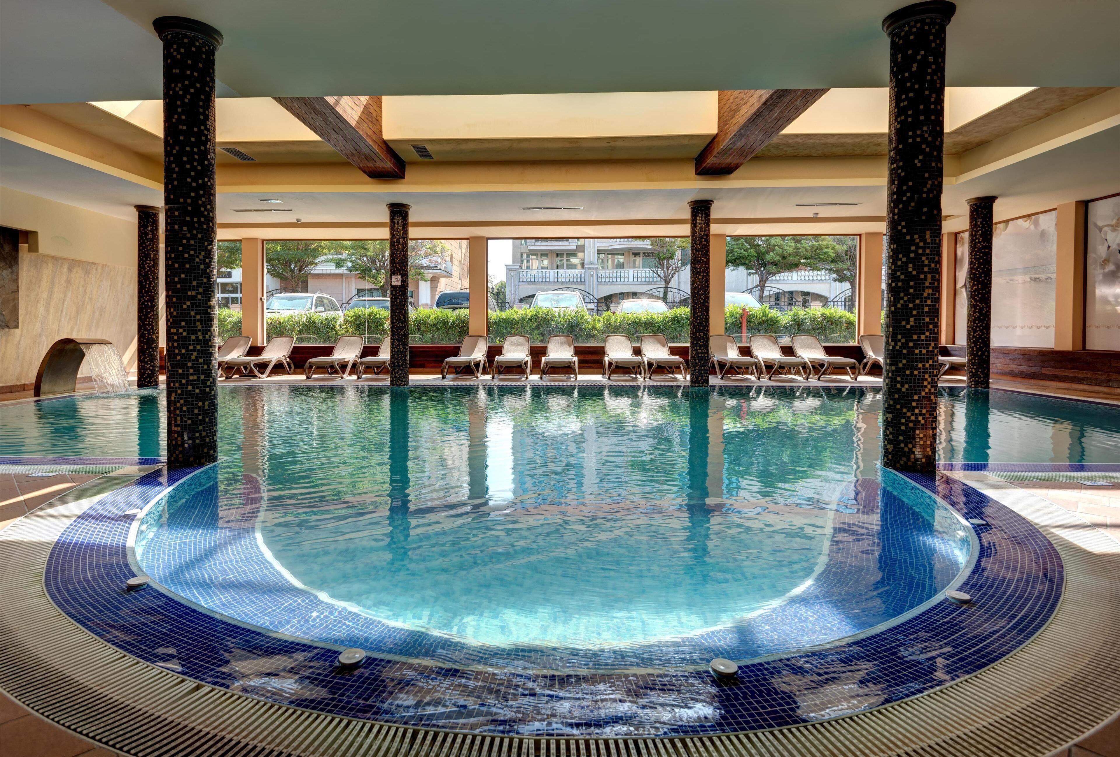 An indoor swimming pool with a tiled edge, lounge chairs, and pillars.