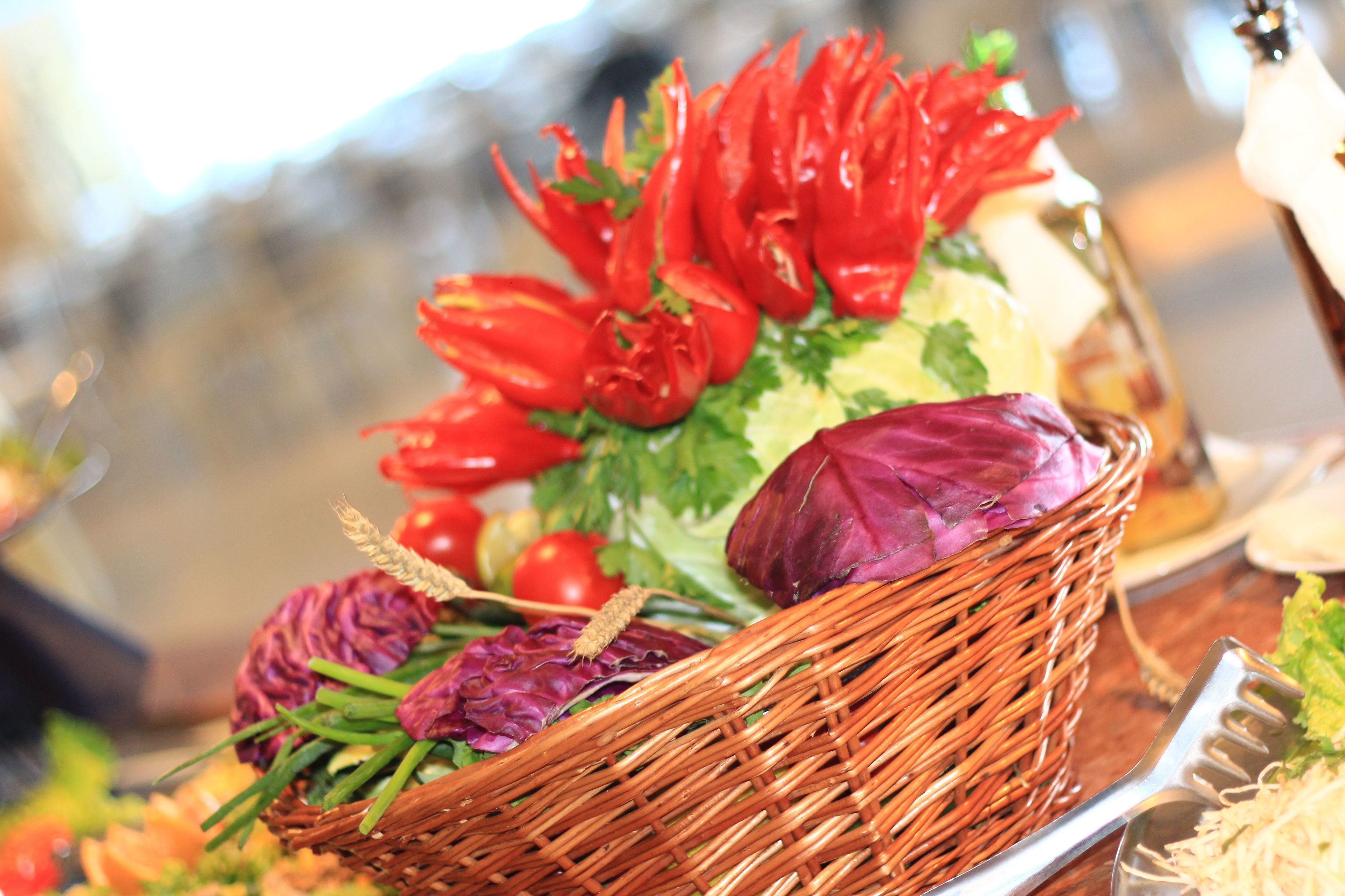 A wicker basket filled with various vegetables and herbs, including red peppers and lettuce, placed on a table.