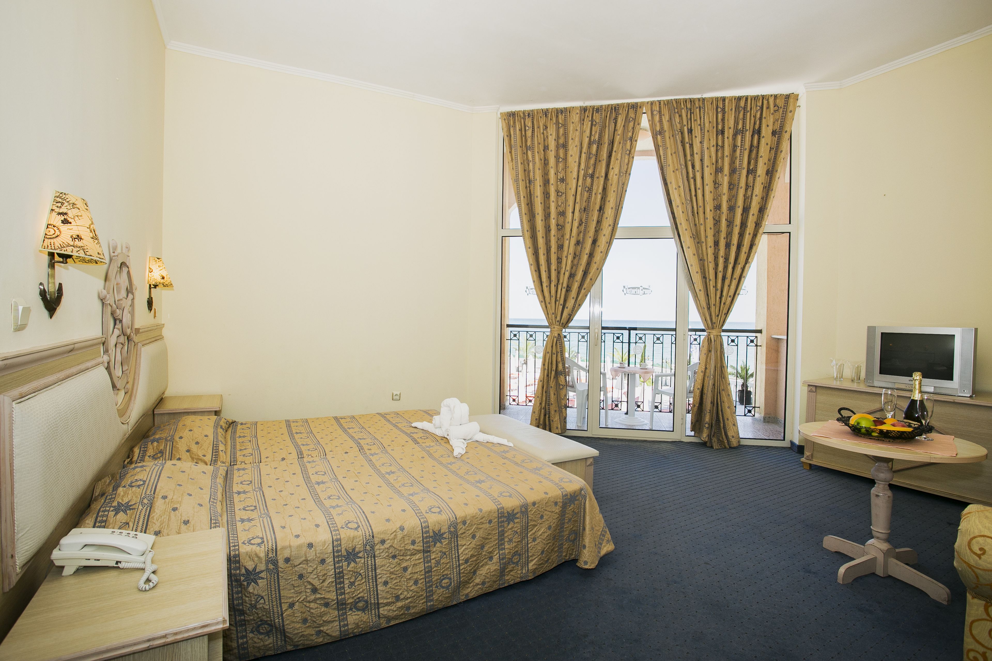 A hotel room with a bed, a desk with a telephone, a TV, and a balcony with curtains.