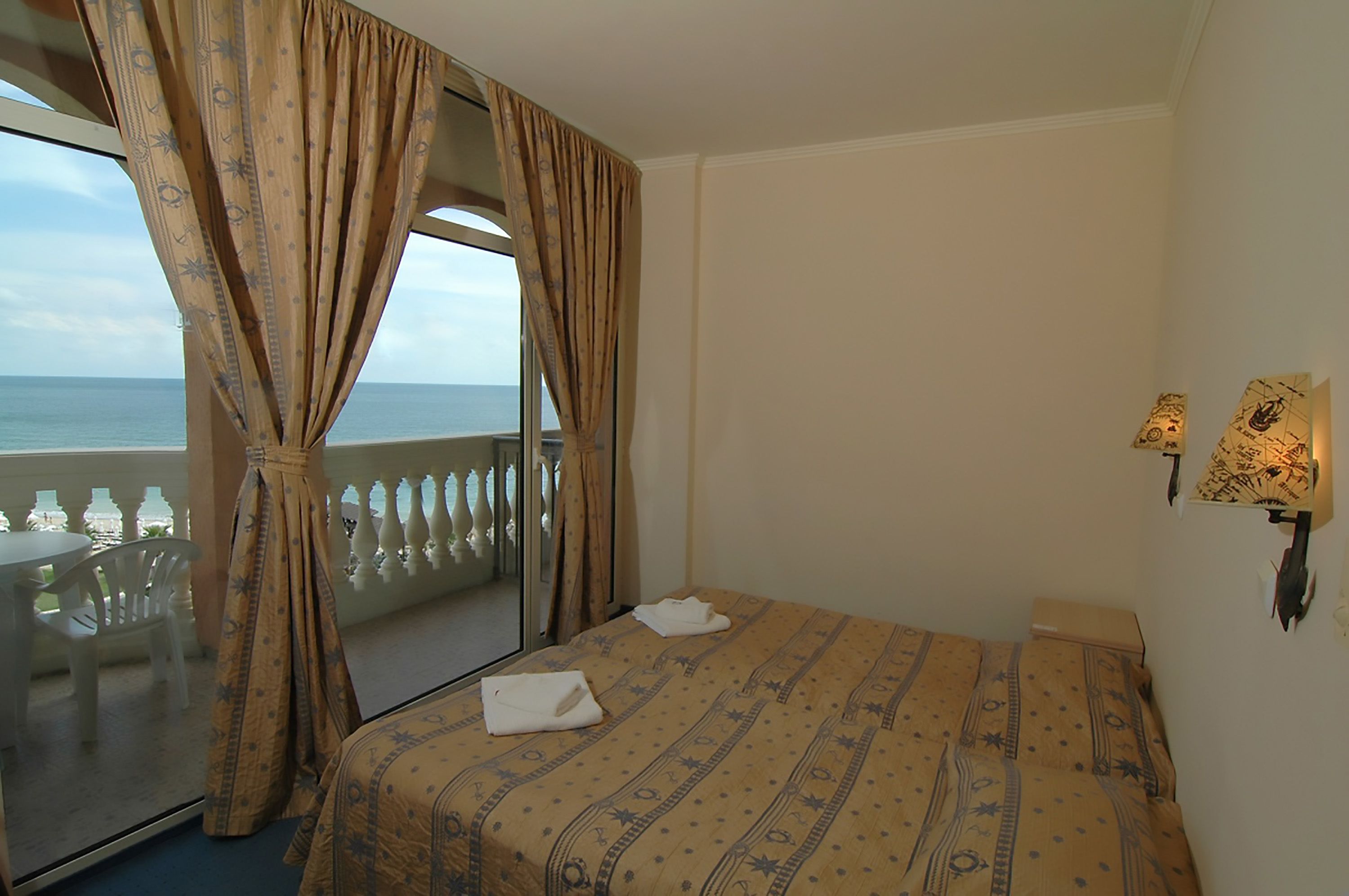 Quarto de hotel com cama, janela com cortinas, varanda com cadeiras e luminária na parede.