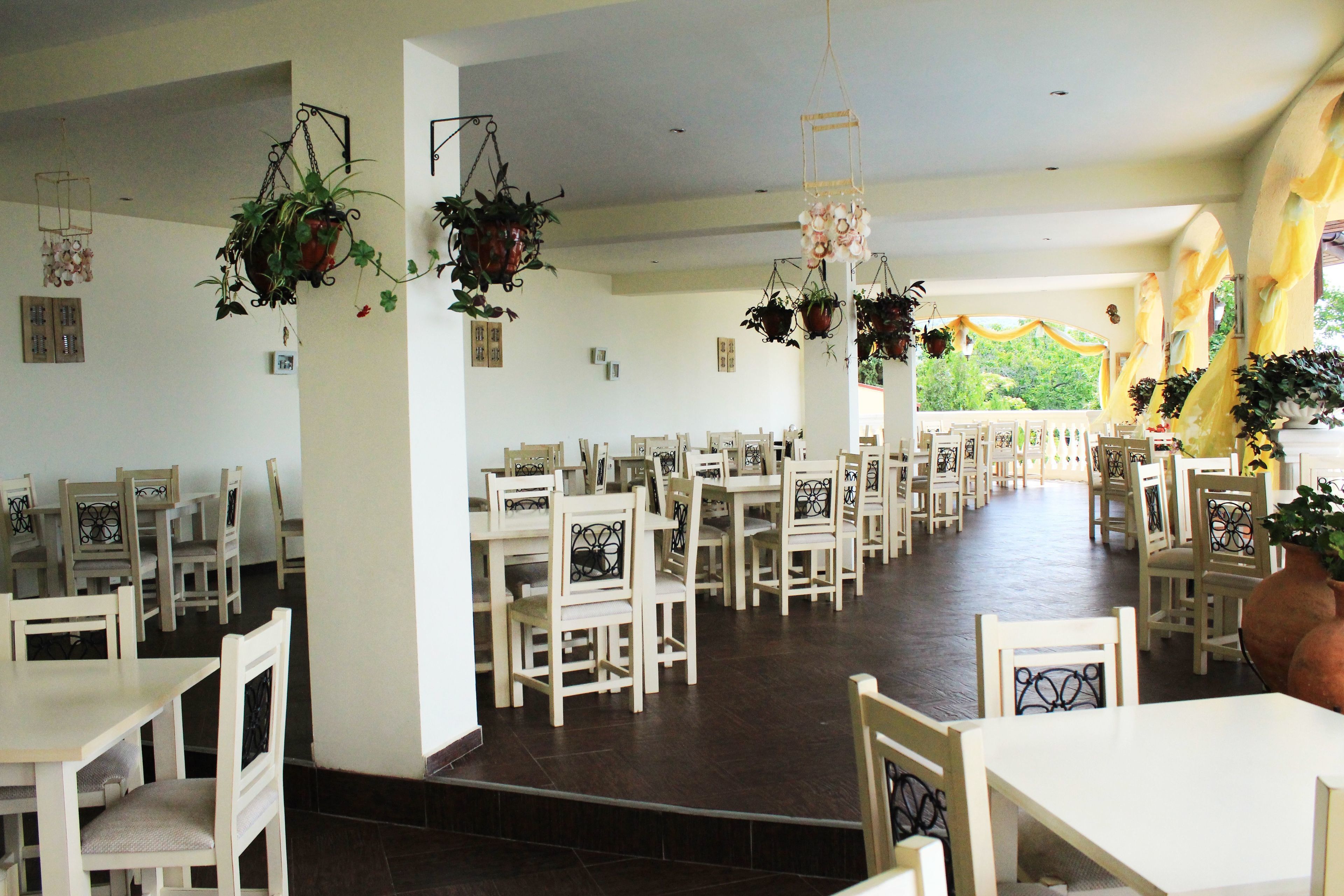 Un comedor amplio con mesas y sillas blancas, plantas colgantes y vistas al exterior.