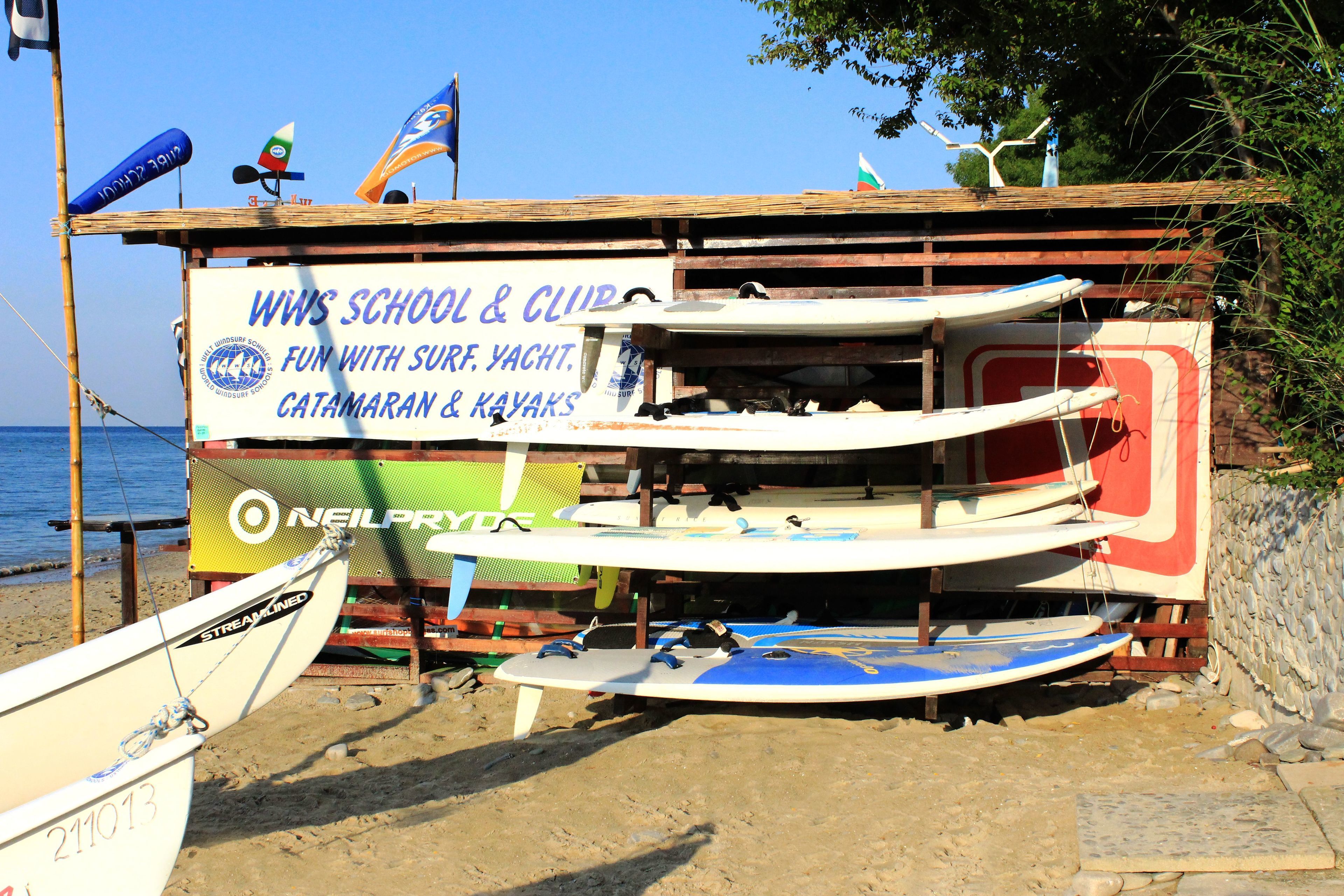 En bygning ved stranden med et skilt, der reklamerer for en skole og en klub for surfing, sejlbåde, katamaran og kajakaktiviteter.