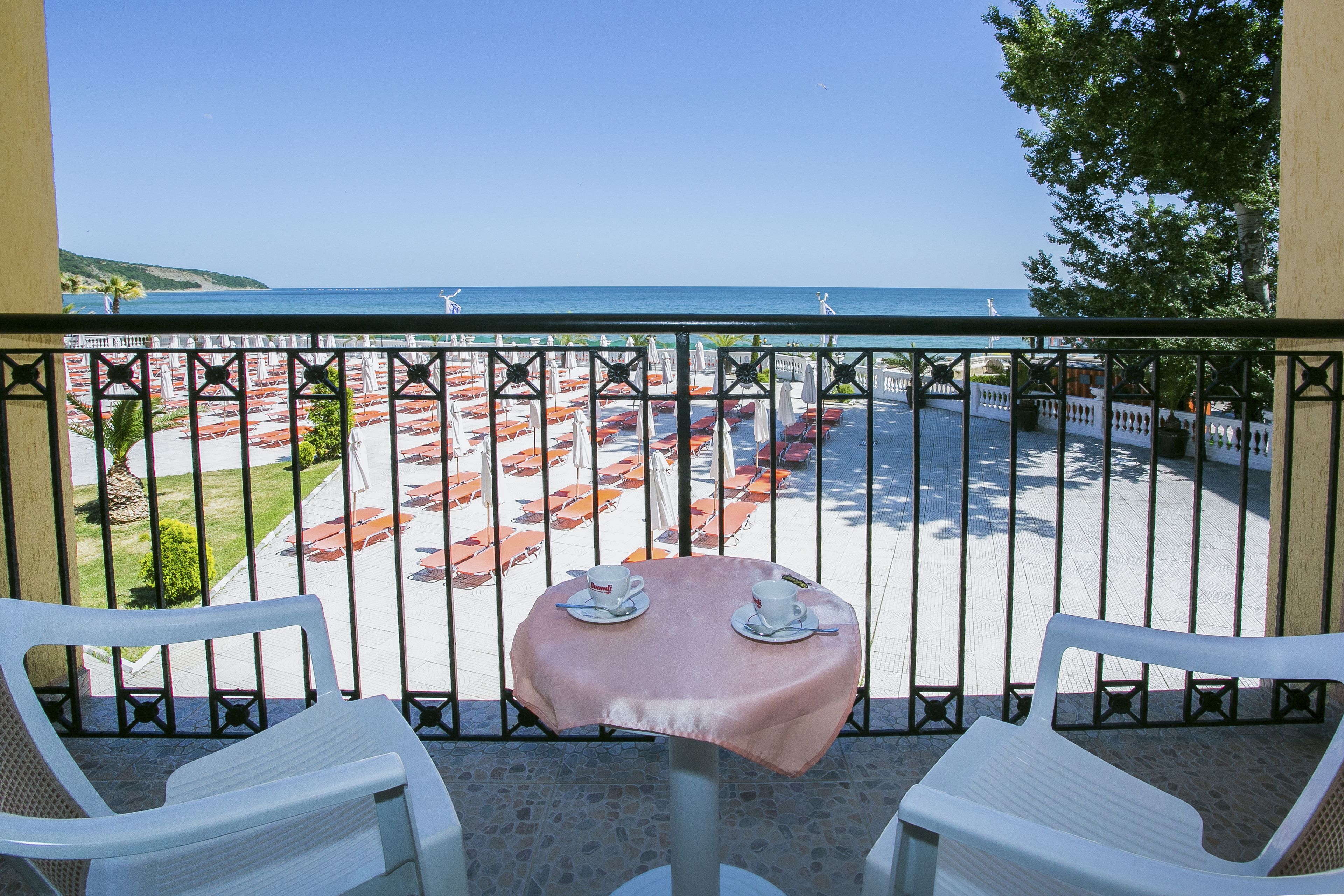 A balcony with a table and two chairs, overlooking a beach with umbrellas and a clear sky.
