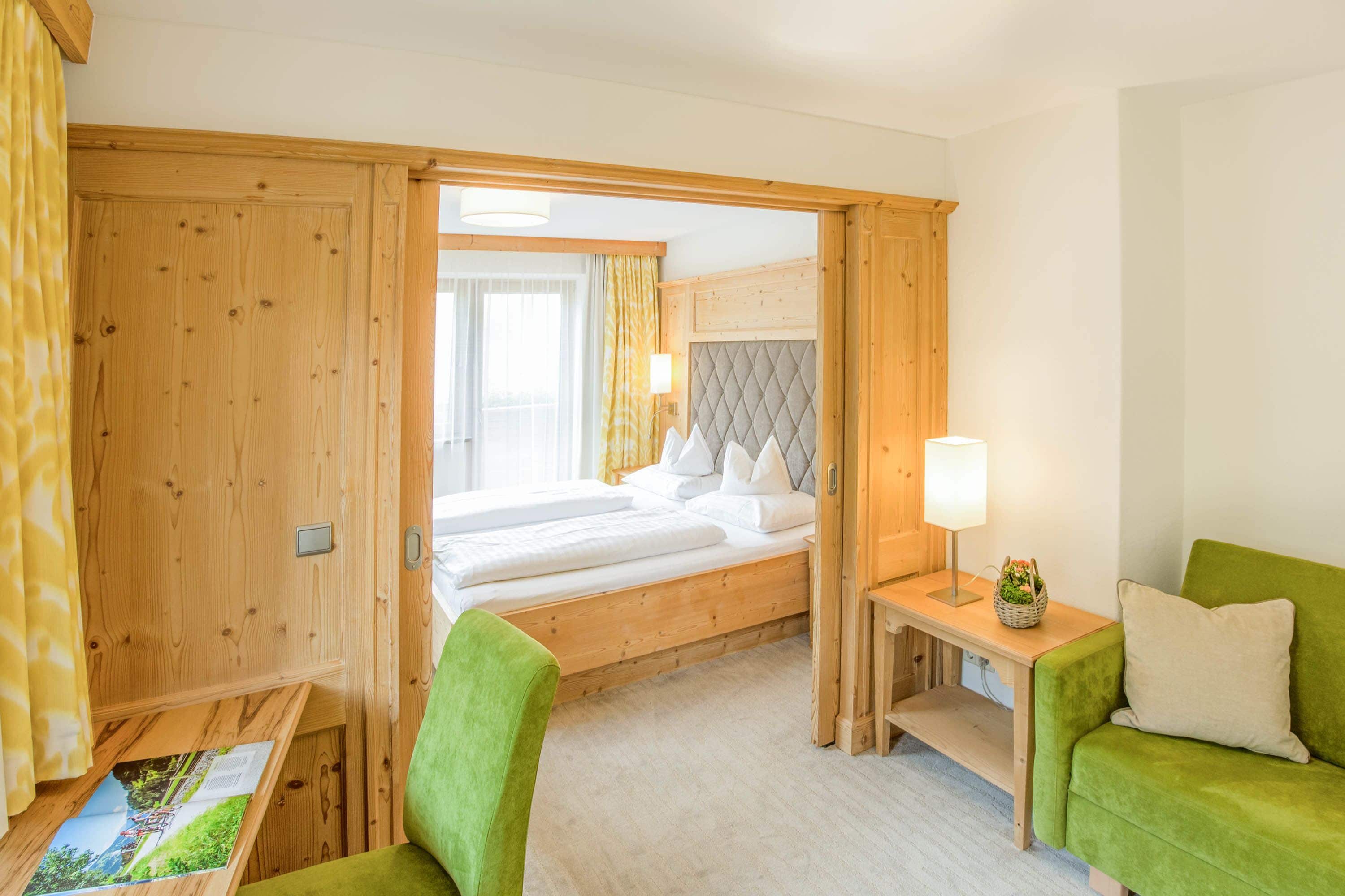Habitación de hotel con cama de madera, un sillón verde, una mesita con lámpara y vista al exterior a través de una ventana.