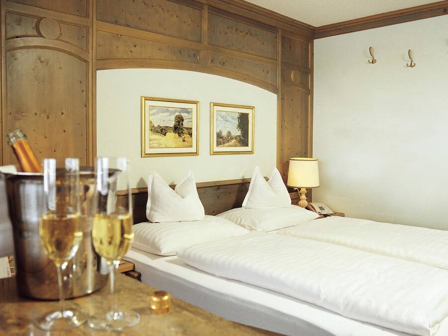 A hotel room with a wooden headboard, two framed pictures on the wall, a bedside table with a lamp, and a bed with white bedding.
