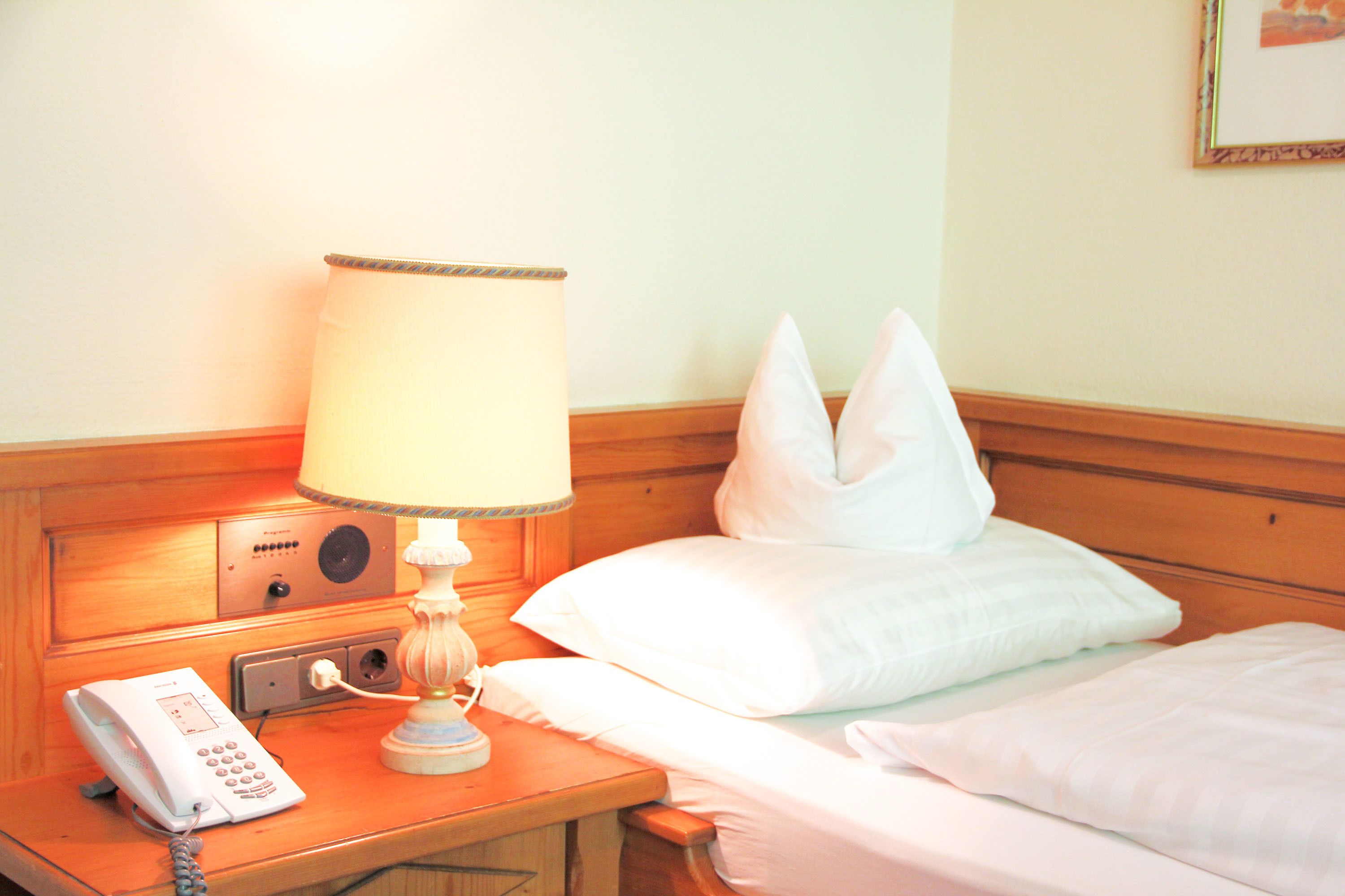 Habitación de hotel con mesita de noche de madera, lámpara blanca, teléfono y cama con ropa de cama blanca.