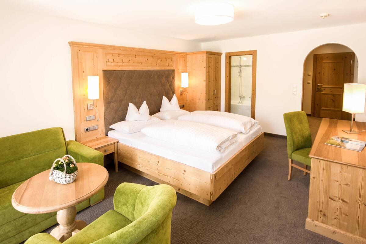 Une chambre d’hôtel avec un lit en bois, des fauteuils verts, une petite table ronde et une armoire en bois.