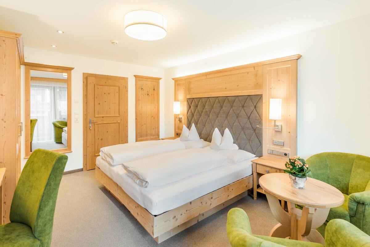 Une chambre d’hôtel avec un grand lit, une tête de lit en bois, deux fauteuils verts, une petite table ronde et une plante en pot.