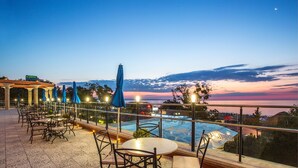An outdoor terrace with tables and chairs, a pool, and a view of the sea at dusk. - Astera Hotel & Spa (Golden Sands)
