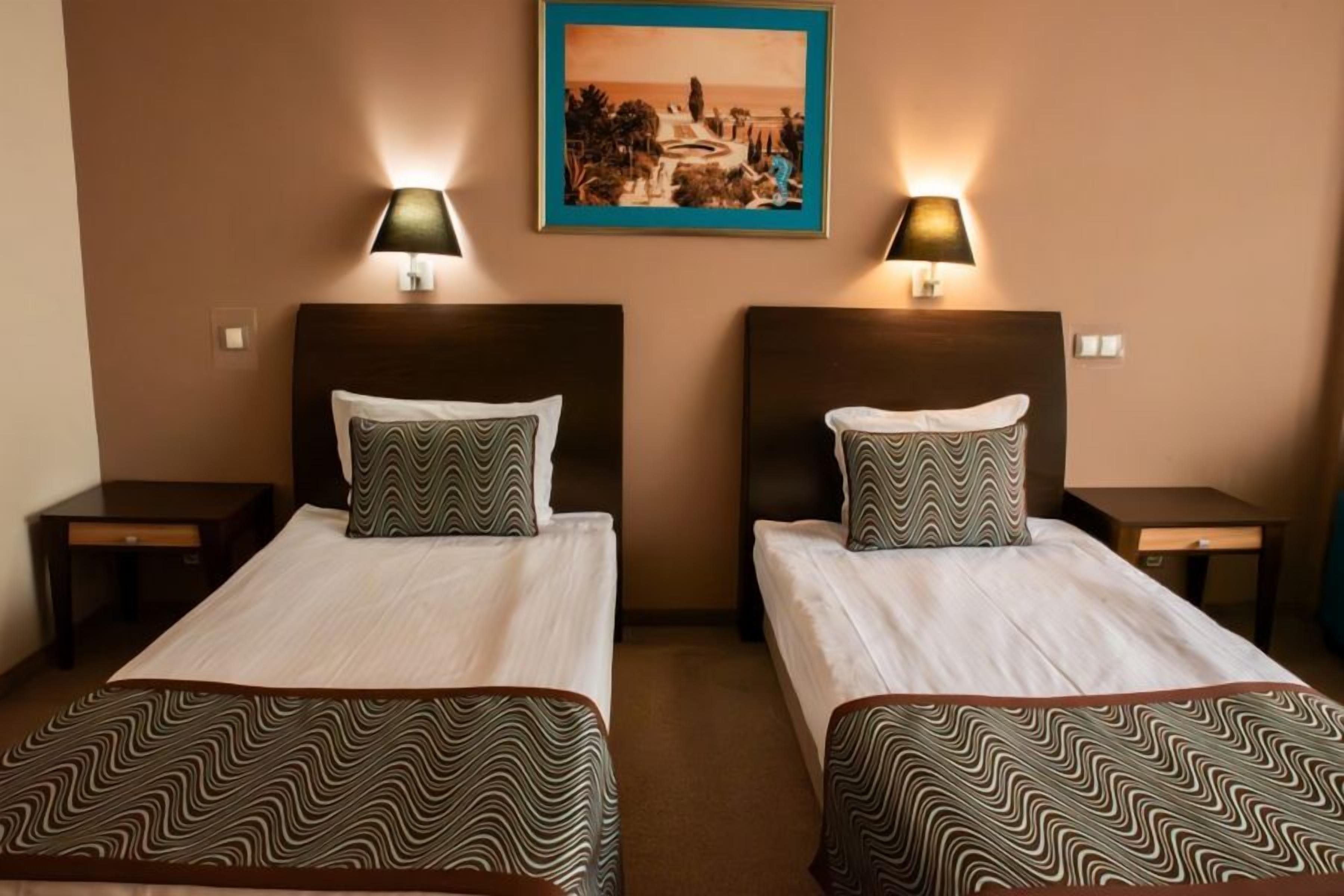 A hotel room with two beds, each with a patterned bedspread and pillow, flanked by nightstands with lamps. A framed picture hangs above the beds.