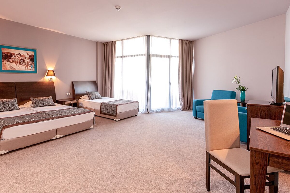 Quarto de hotel com duas camas, uma escrivaninha com um laptop, uma cadeira e uma janela grande com cortinas.