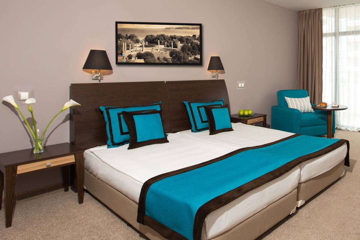 A hotel room with a large bed, two bedside tables, a chair, a small table, and a wall-mounted picture.