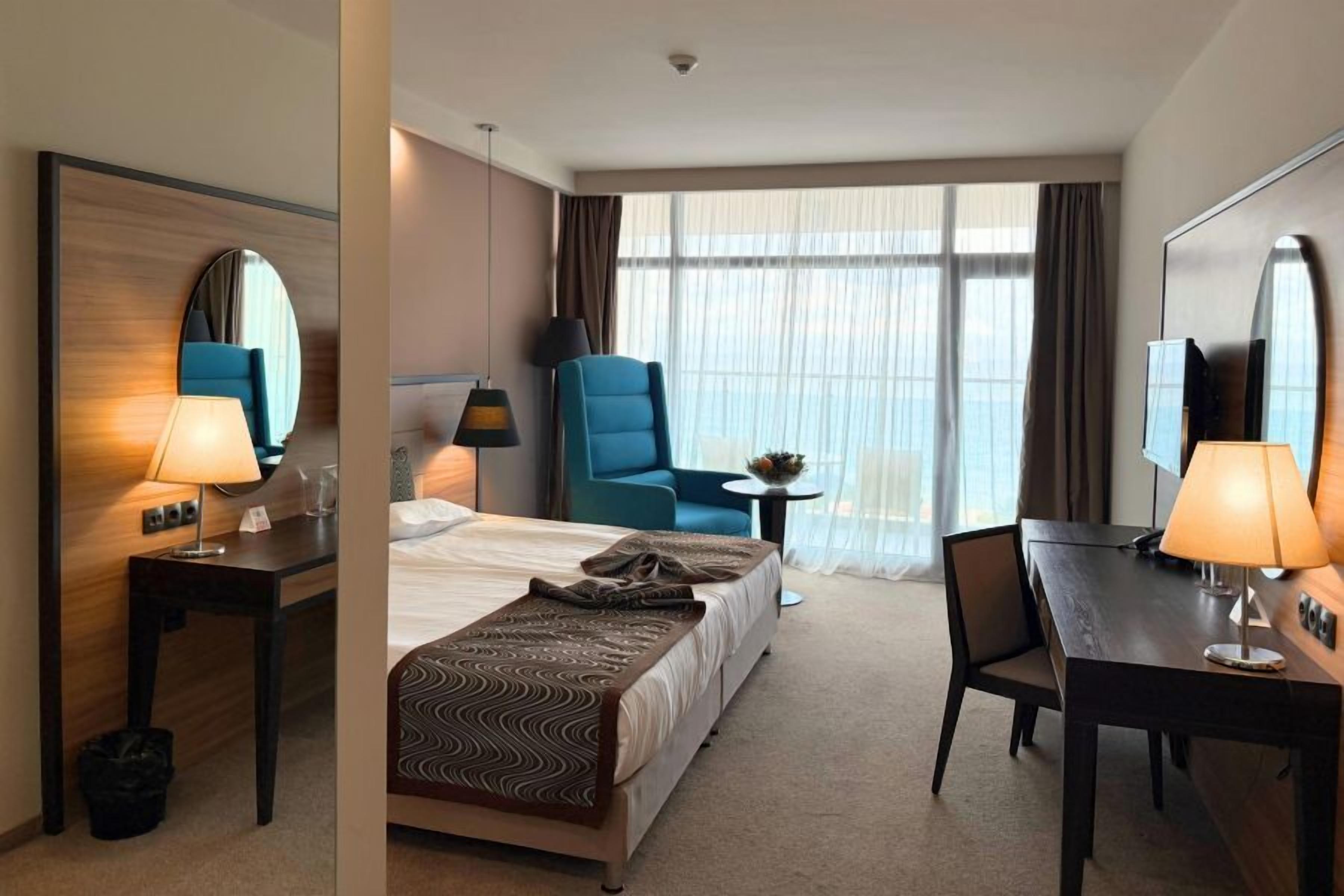 Une chambre d’hôtel comprenant un lit, un bureau, une chaise, une table, une lampe, un miroir et une fenêtre avec des rideaux.
