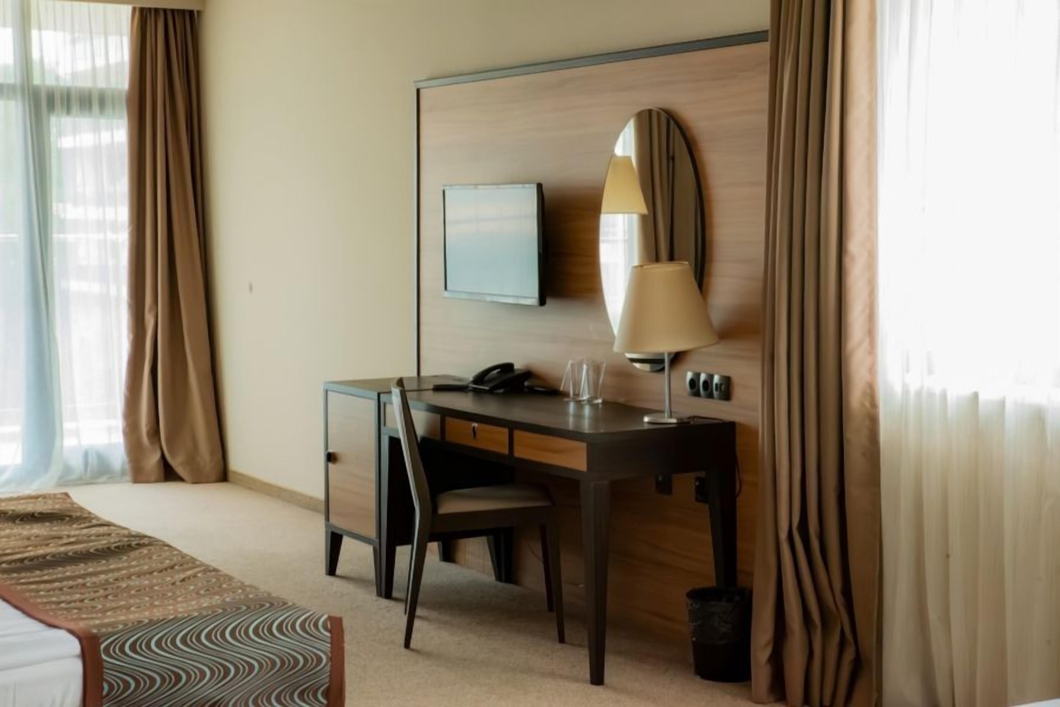 A hotel room with a bed, a desk, a chair, a TV, a lamp, and a waste bin.