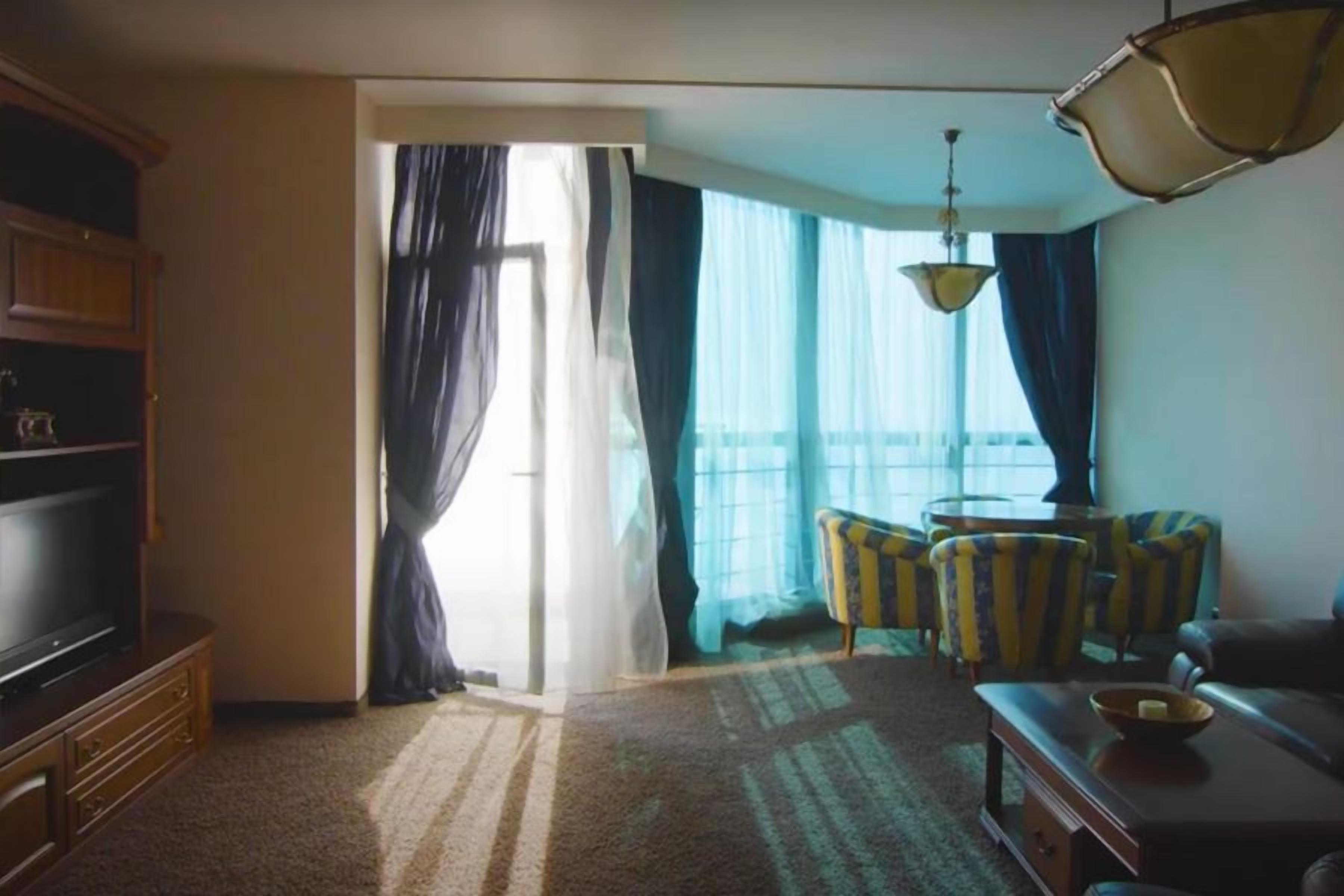 A hotel room with a television, a sofa, striped armchairs, a small table, and a view through a window with curtains.