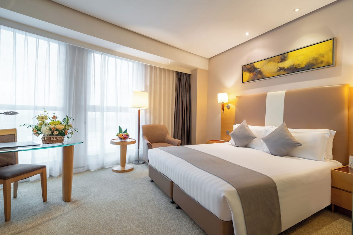 A hotel room with a large bed, a desk with a chair, a small table with a lamp, and a window with curtains.
