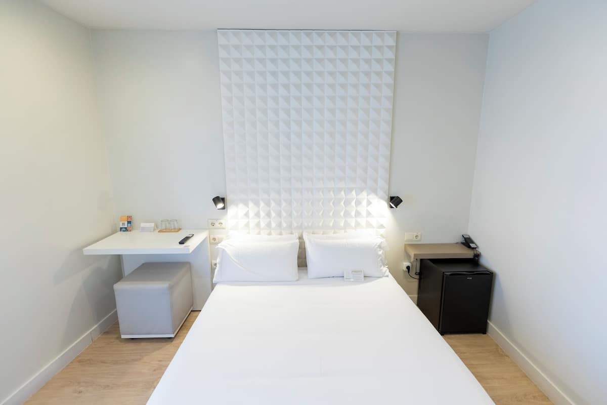 A minimalist hotel room with a bed, bedside tables, a desk, and a small black refrigerator.