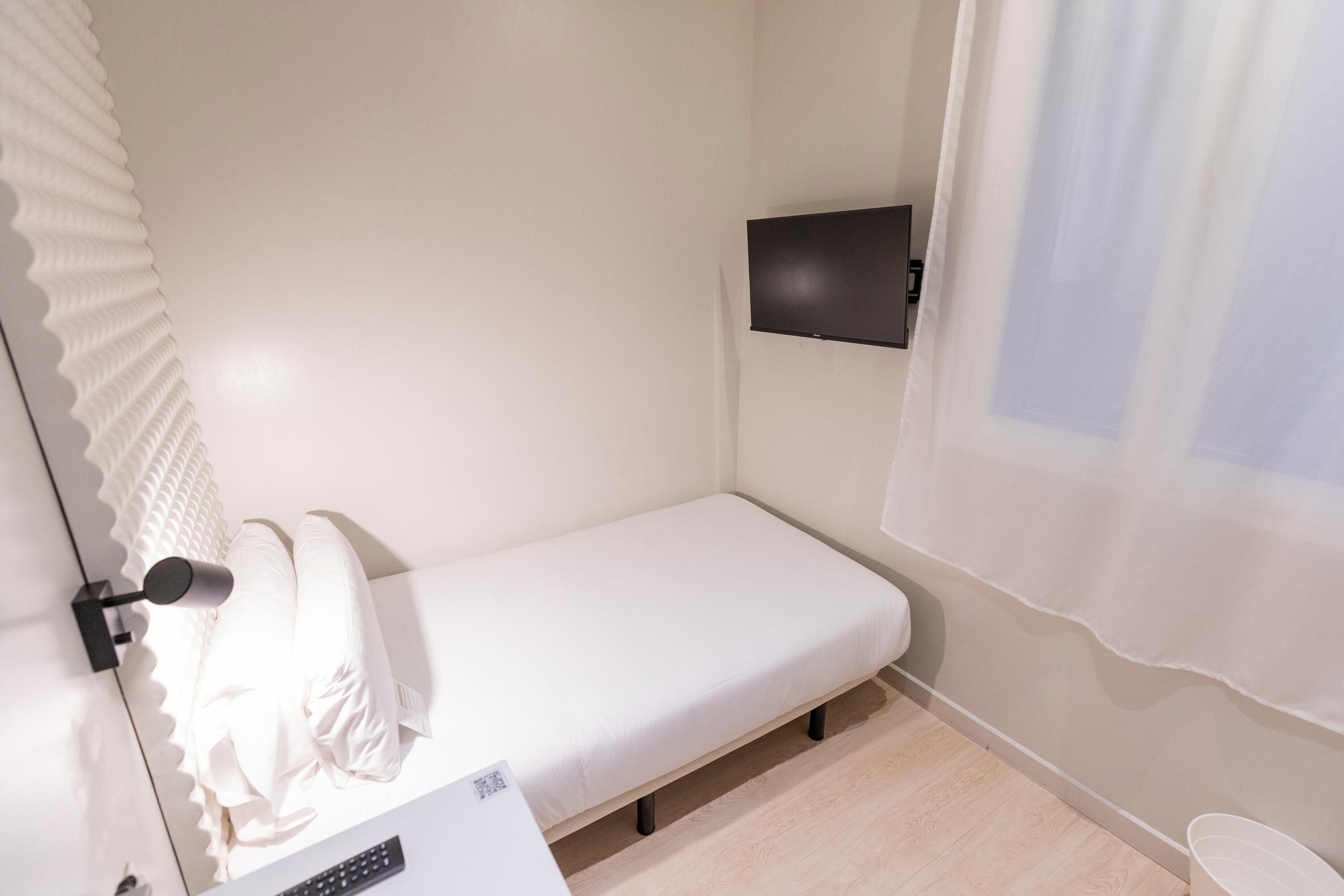 A small, single-bed room with a white mattress, a bedside table, a mounted TV, and a window with sheer curtains.