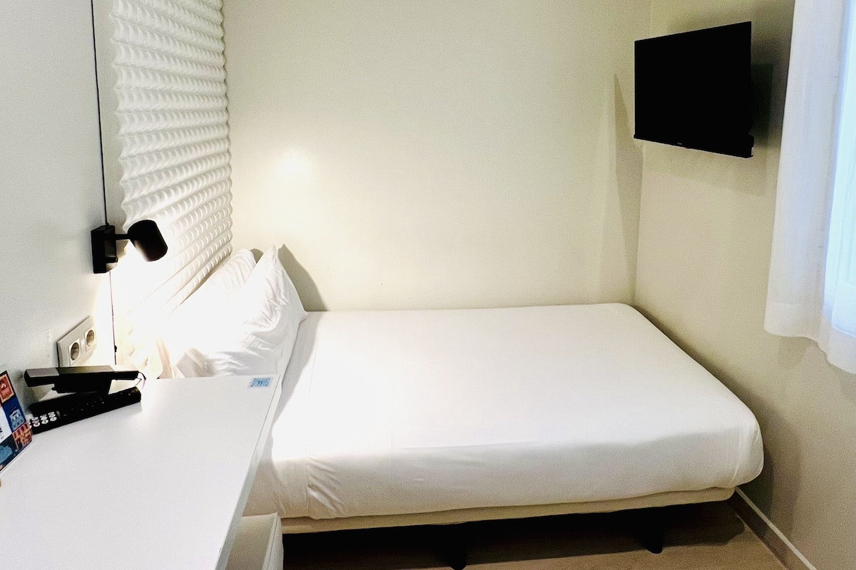 A small, single-bed room with a white bed, a bedside table with a lamp, a flat-screen TV mounted on the wall, and a window with blinds.