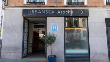 Exterior del hotel URBANSEA Atocha 113 con una planta grande en maceta, una entrada visible y una vista clara del interior a través de la fachada de vidrio.