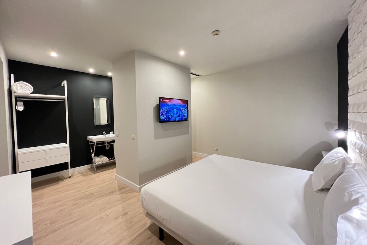 A modern hotel room with a large bed, a built-in TV, a small bathroom with a sink and mirror, and a walk-in closet.