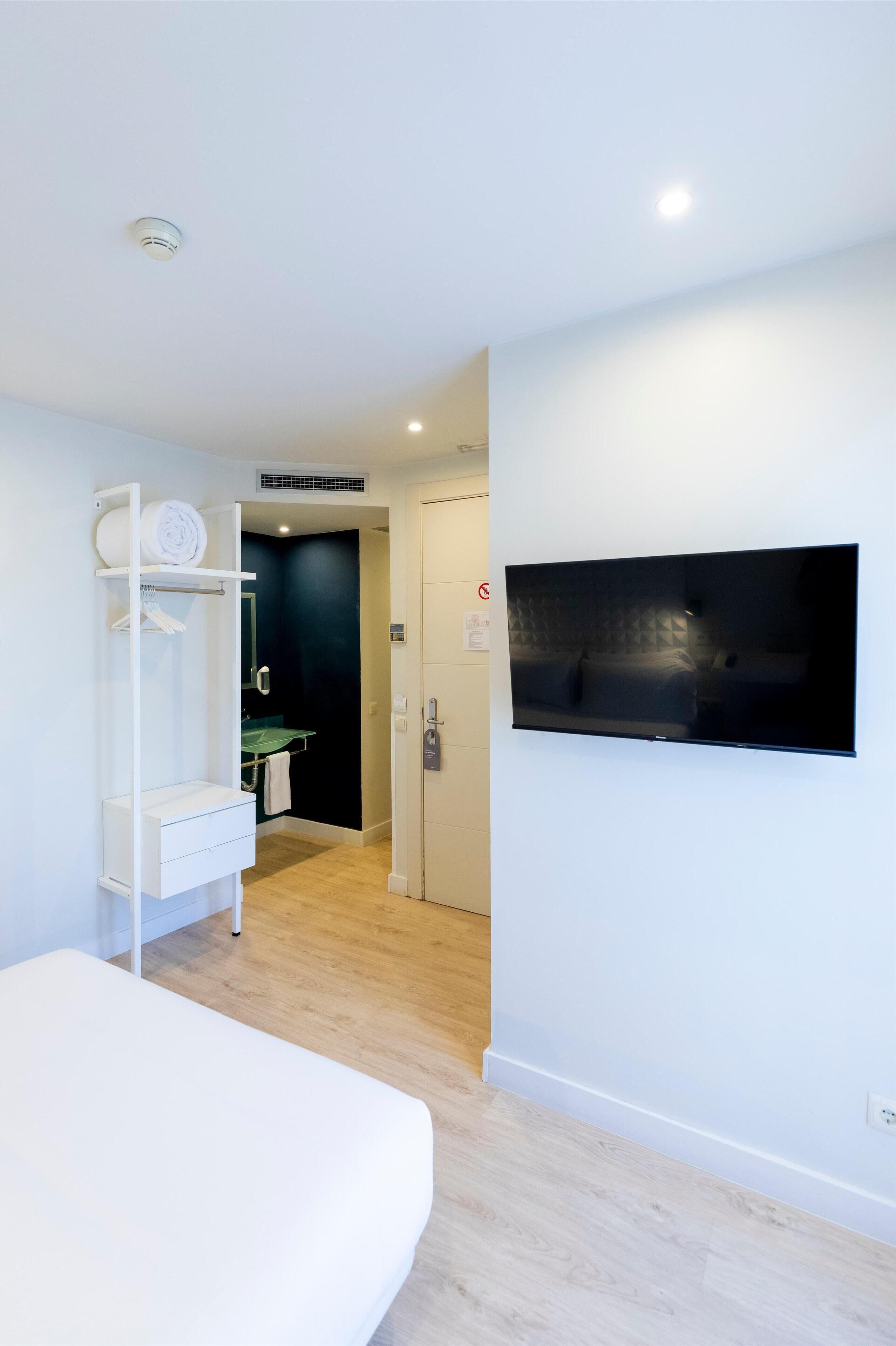 A modern hotel room with a flat-screen TV mounted on the wall, a white wardrobe with a mirror, and a wooden floor.