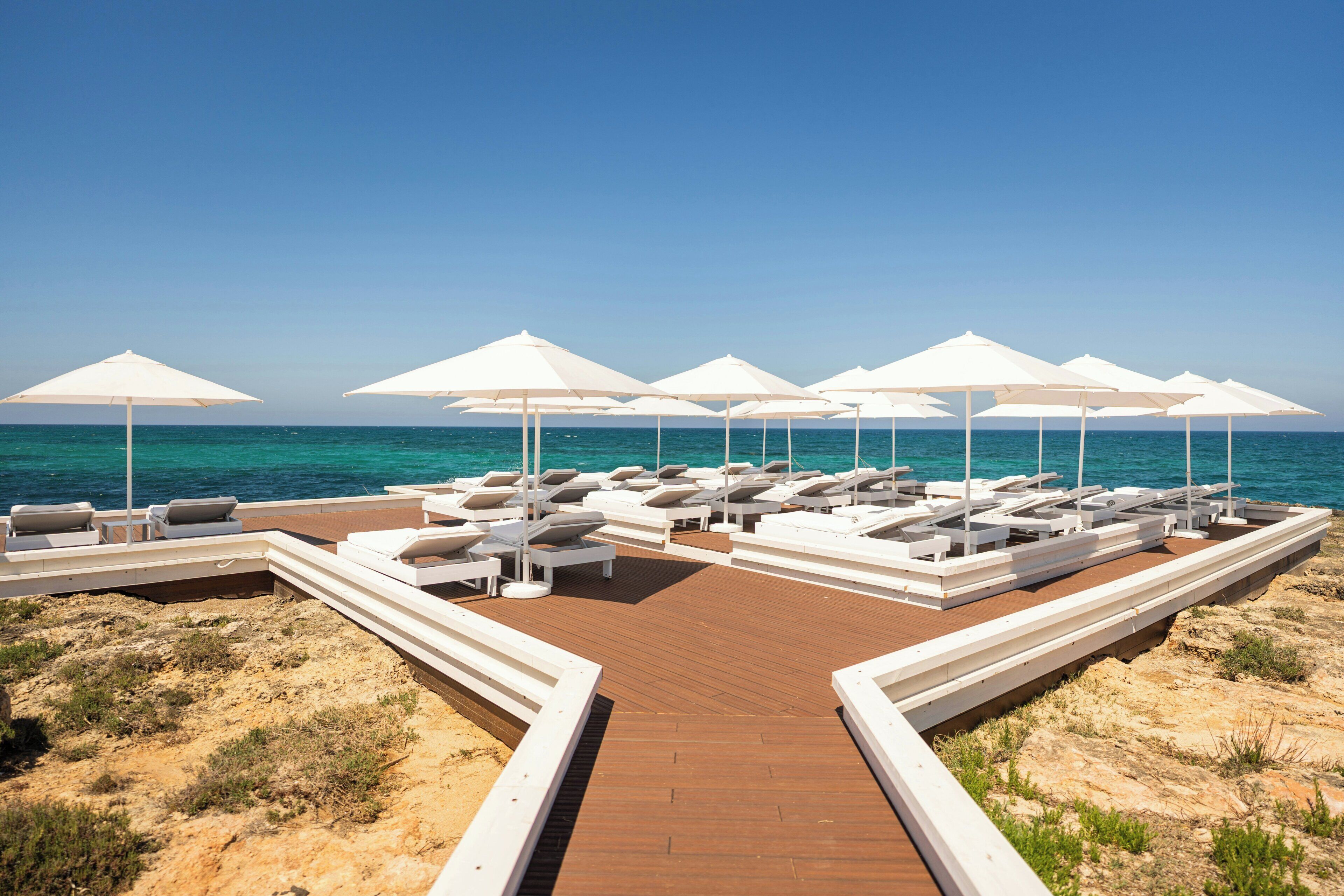 Un espace de détente aménagé sur la plage, avec des parasols blancs et des chaises longues, une terrasse en bois et une vue dégagée sur la mer.