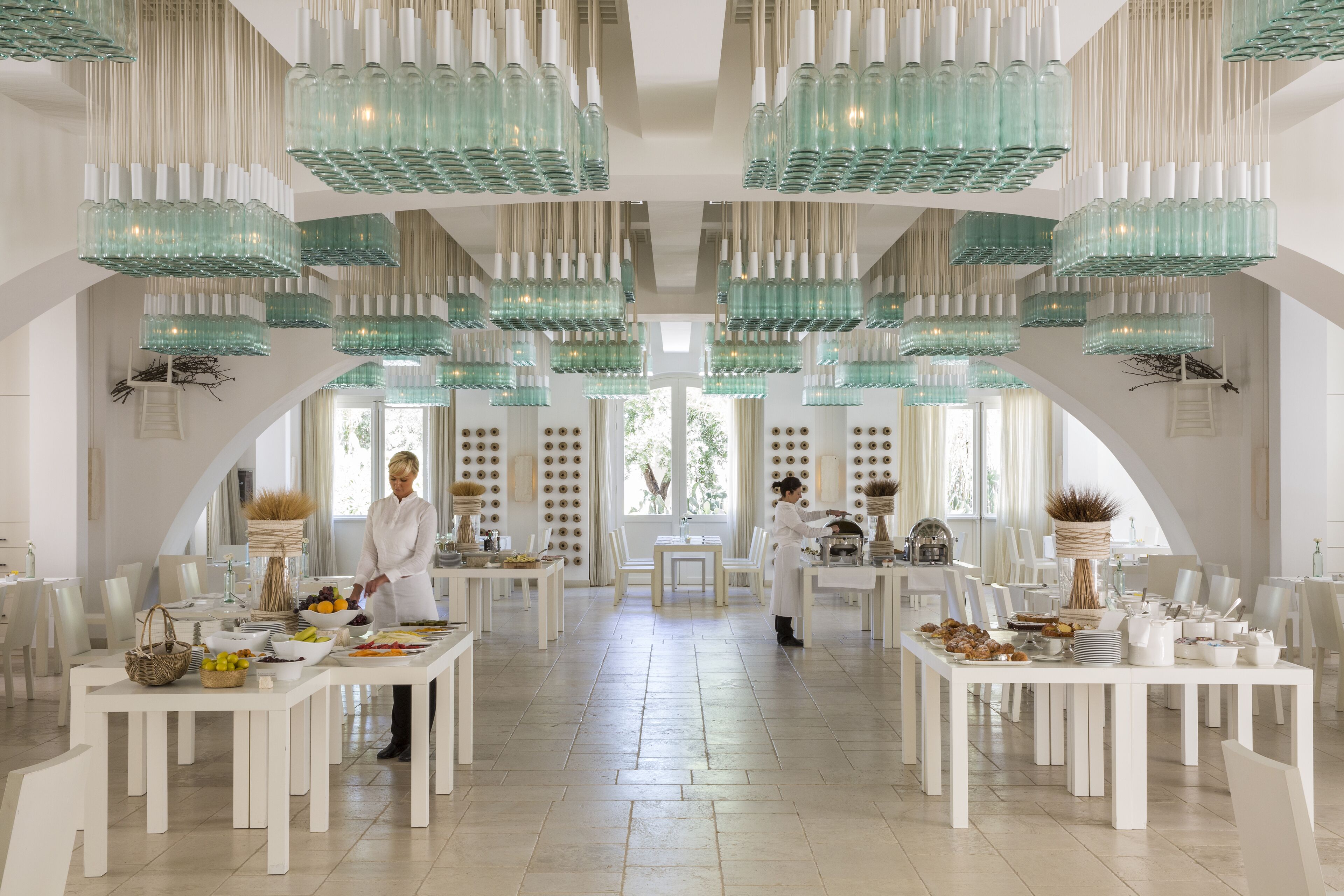 Un comedor amplio con mesas y sillas blancas, un área central de servicio y lámparas decorativas colgantes.