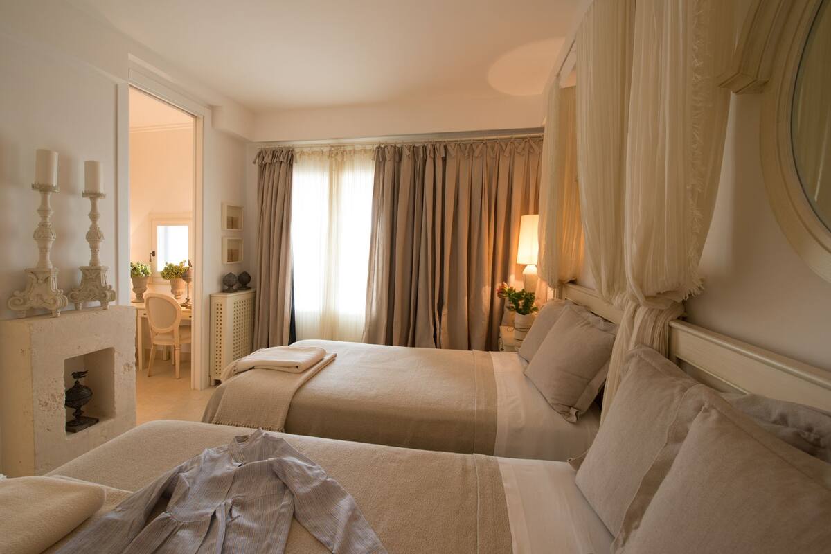 A hotel room with two beds, a fireplace, a desk with a chair, and a large window with curtains.
