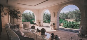 Um pátio banhado pelo sol, com mesa de vidro, sofá e vista para um jardim através de arcos.